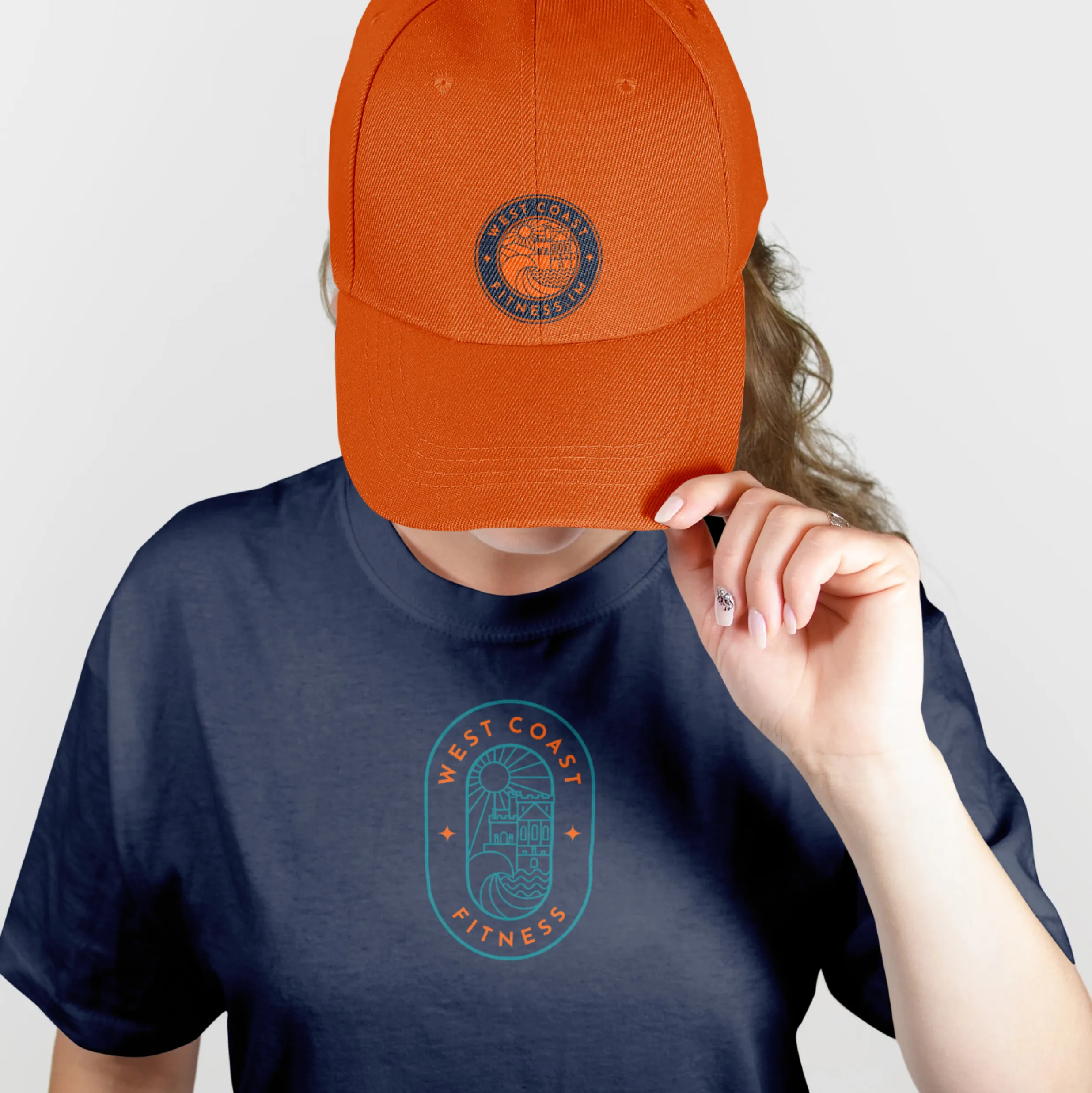 Woman wearing a navy West Coast Fitness branded t-shirt and orange branded cap against a light grey background